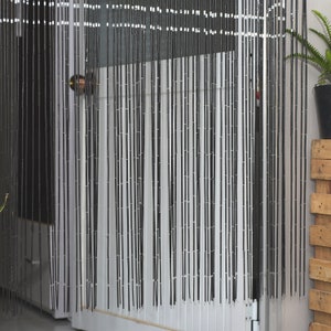 May include: Black beaded curtain hanging in a doorway. The curtain is made of numerous vertical strands of black beads, creating a textured, semi-opaque barrier. The beads are strung together with white string. A door and a potted plant are visible behind the curtain.