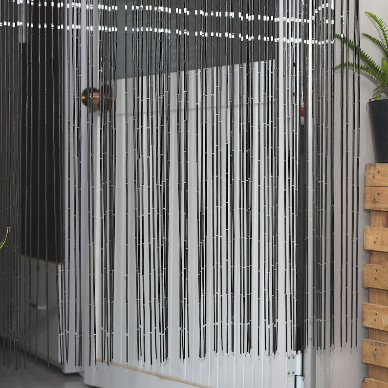 May include: Black beaded curtain hanging in a doorway. The curtain is made of numerous vertical strands of black beads, creating a textured, semi-opaque barrier. The beads are strung together with white string. A door and a potted plant are visible behind the curtain.