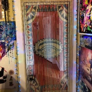 May include: A white beaded door curtain with a red, white, and blue patterned tapestry hanging behind it. The curtain is strung with white beads and has a fringe at the bottom. The tapestry has a geometric design with a large eye in the center.