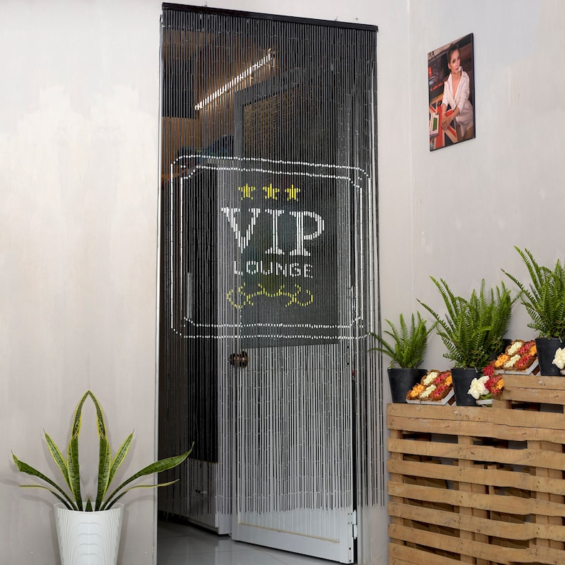 May include: Black beaded door curtain with a white VIP LOUNGE sign. The sign features three gold stars and a decorative yellow design. The curtain hangs in a doorway, with a glimpse of the interior visible. A potted plant is in the foreground.