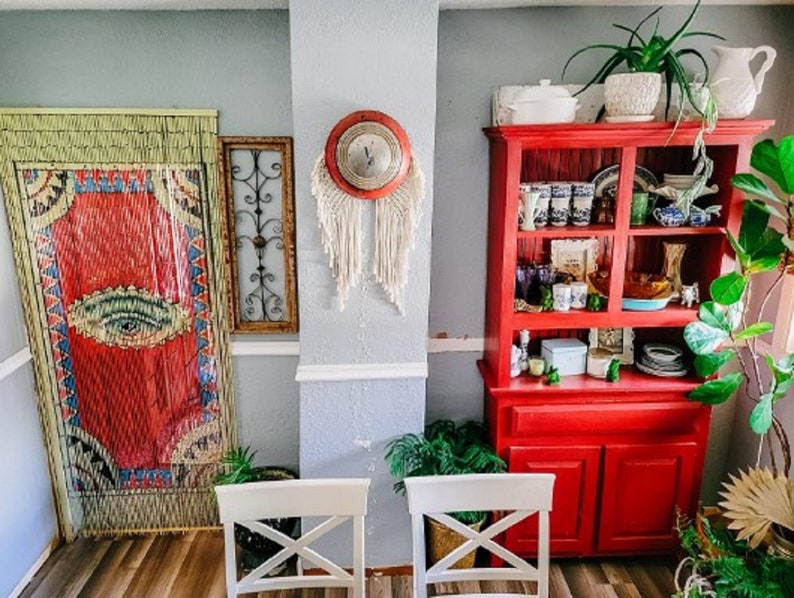 May include: A red hutch with glass doors and shelves filled with decorative items. The hutch has a cabinet with two doors at the bottom. There is a large green plant in the corner of the room.