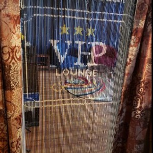 May include: A bamboo beaded curtain with a sign that reads "VIP LOUNGE". The sign is blue with gold stars and white and purple lettering. The curtain is flanked by patterned brown curtains.