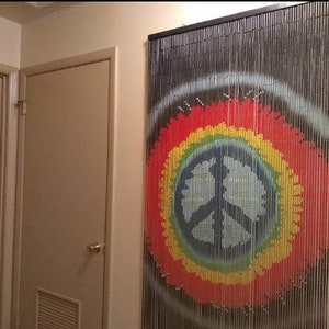May include: A beaded curtain with a peace sign design. The curtain features a black frame and strands, with a colorful peace sign in red, yellow, green, and blue. The curtain hangs on a beige wall.