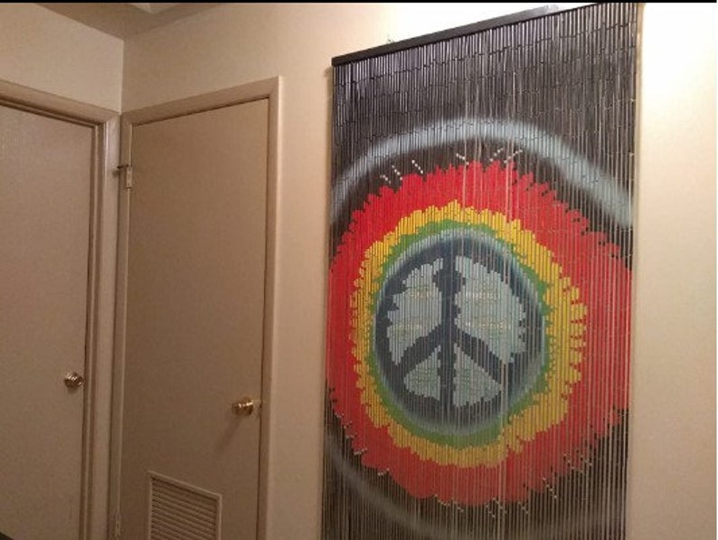 May include: A beaded curtain with a peace sign design. The curtain features a black frame and strands, with a colorful peace sign in red, yellow, green, and blue. The curtain hangs on a beige wall.