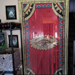 May include: A red and yellow beaded curtain with a large eye design in the center. The curtain is made of wooden beads and has a geometric pattern on the sides.