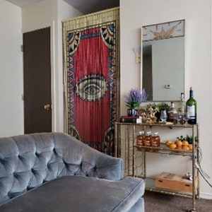 May include: A blue velvet tufted sofa sits in front of a red and blue beaded curtain with an eye design. A gold metal bar cart with glass shelves holds bottles and glasses. The bar cart is on a beige carpet.