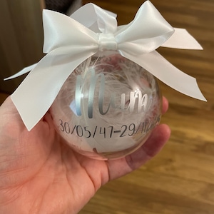 May include: A clear spherical bauble filled with white feathers, featuring a white satin ribbon bow at the top. The surface is printed with the word Mum in a silver script font, followed by the date 30/05/47-29/11/22 in a silver sans-serif font.