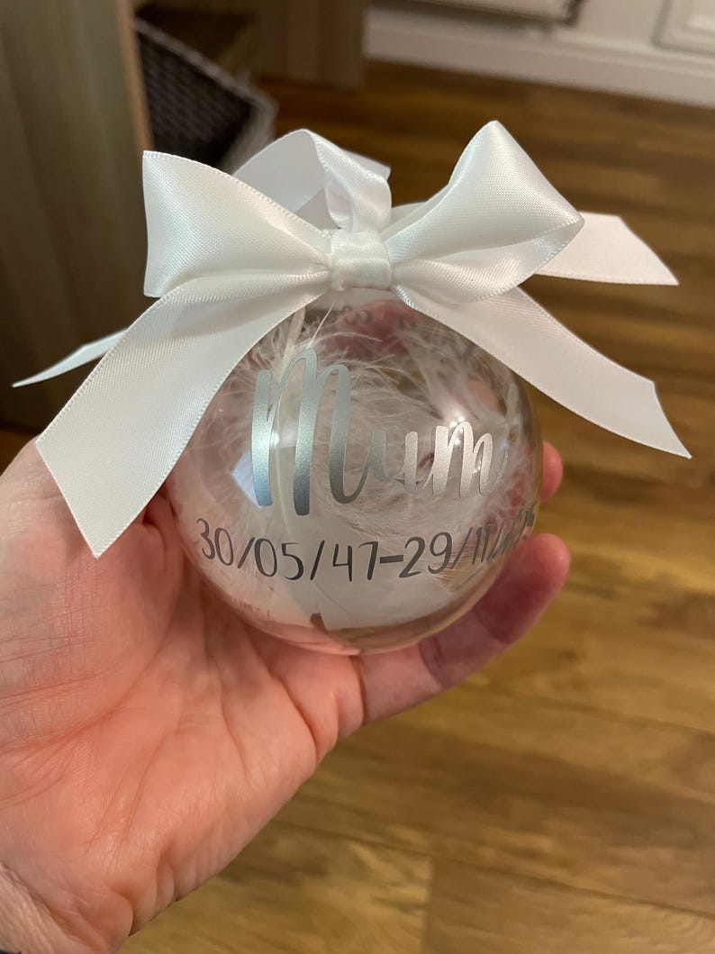 May include: A clear spherical bauble filled with white feathers, featuring a white satin ribbon bow at the top. The surface is printed with the word Mum in a silver script font, followed by the date 30/05/47-29/11/22 in a silver sans-serif font.