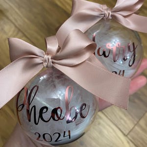 May include: Two clear spherical baubles filled with white feathers, each topped with a dusty pink satin ribbon bow. The front of one bauble features the name Phoebe and the year 2024 in metallic rose gold script and block lettering.