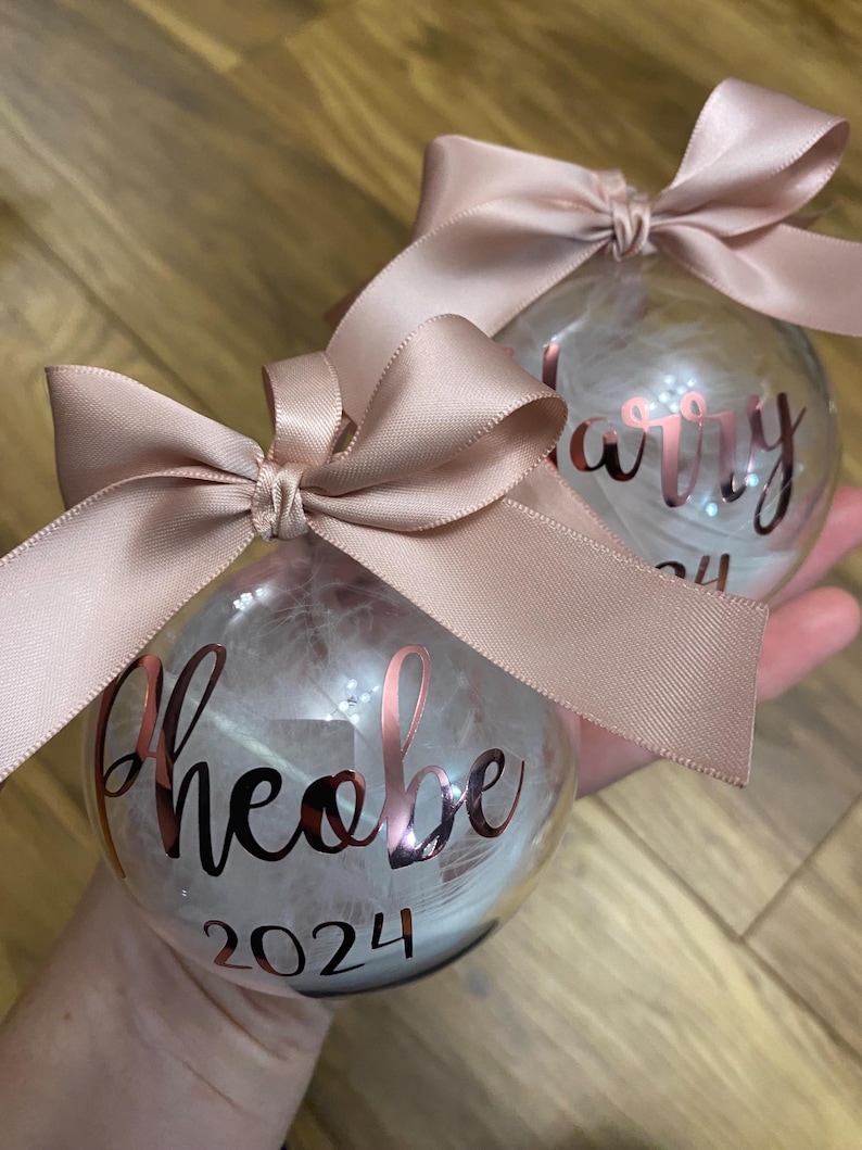 May include: Two clear spherical baubles filled with white feathers, each topped with a dusty pink satin ribbon bow. The front of one bauble features the name Phoebe and the year 2024 in metallic rose gold script and block lettering.