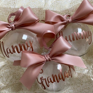 May include: Three clear spherical baubles filled with white feathers, each topped with a large dusty rose satin ribbon bow. The baubles feature metallic rose gold lettering that reads Nanny, Mummy, and Grandad in a cursive script font.