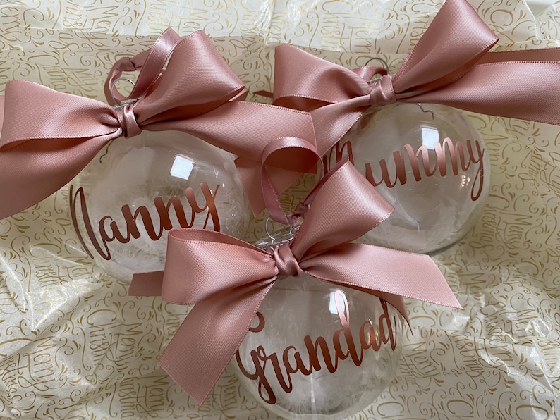 May include: Three clear spherical baubles filled with white feathers, each topped with a large dusty rose satin ribbon bow. The baubles feature metallic rose gold lettering that reads Nanny, Mummy, and Grandad in a cursive script font.