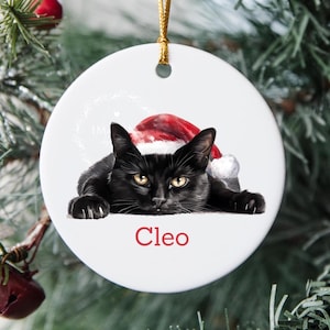 May include: A white ceramic Christmas bauble with a black cat wearing a Santa hat. The cat's name, "Cleo," is printed in red at the bottom. The bauble hangs from a gold string and is surrounded by Christmas tree branches.