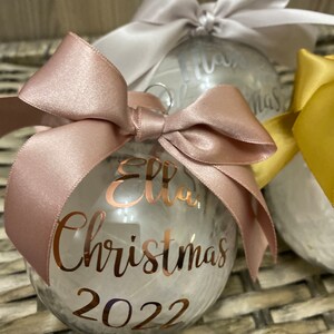 May include: A clear spherical Christmas bauble filled with white feathers, topped with a dusty rose satin ribbon bow. The front features rose gold metallic lettering that reads Ella, Christmas, and 2022 in a cursive and print font combination.