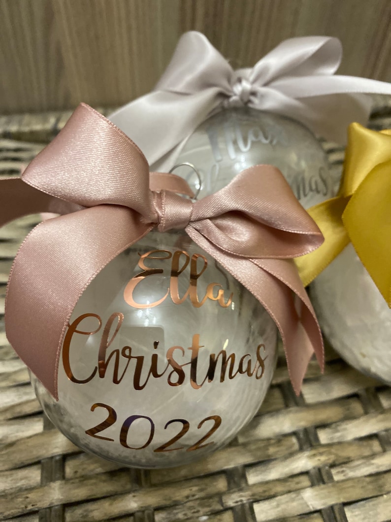 May include: A clear spherical Christmas bauble filled with white feathers, topped with a dusty rose satin ribbon bow. The front features rose gold metallic lettering that reads Ella, Christmas, and 2022 in a cursive and print font combination.