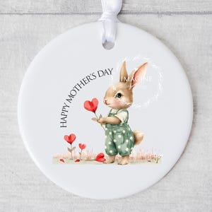 May include: White ceramic ornament featuring a watercolour illustration of a bunny holding a red heart-shaped flower. The text "HAPPY MOTHER'S DAY" is written in a circle around the bunny. The bunny is wearing a green and white polka dot overall.