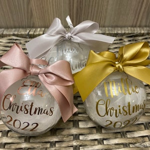 May include: Three clear spherical Christmas baubles filled with white material. Each bauble features a satin ribbon bow on top and personalised text in metallic script. The baubles display the names Ella, Max, and Millie with the text Christmas 2022.