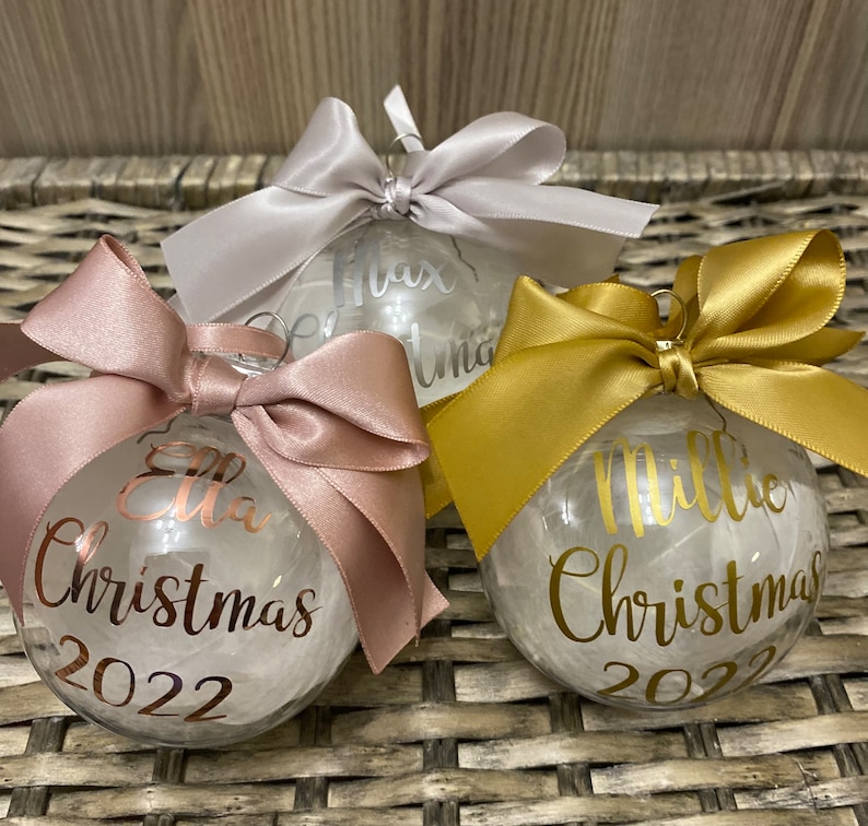 May include: Three clear spherical Christmas baubles filled with white material. Each bauble features a satin ribbon bow on top and personalised text in metallic script. The baubles display the names Ella, Max, and Millie with the text Christmas 2022.