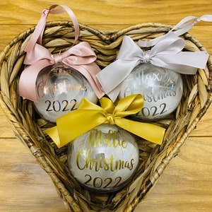 May include: Three clear spherical Christmas baubles filled with white feathers. Each bauble features a satin ribbon bow and custom text in metallic vinyl: Ella Christmas 2022 in rose gold, Max Christmas 2022 in silver, and Millie Christmas 2022 in gold.