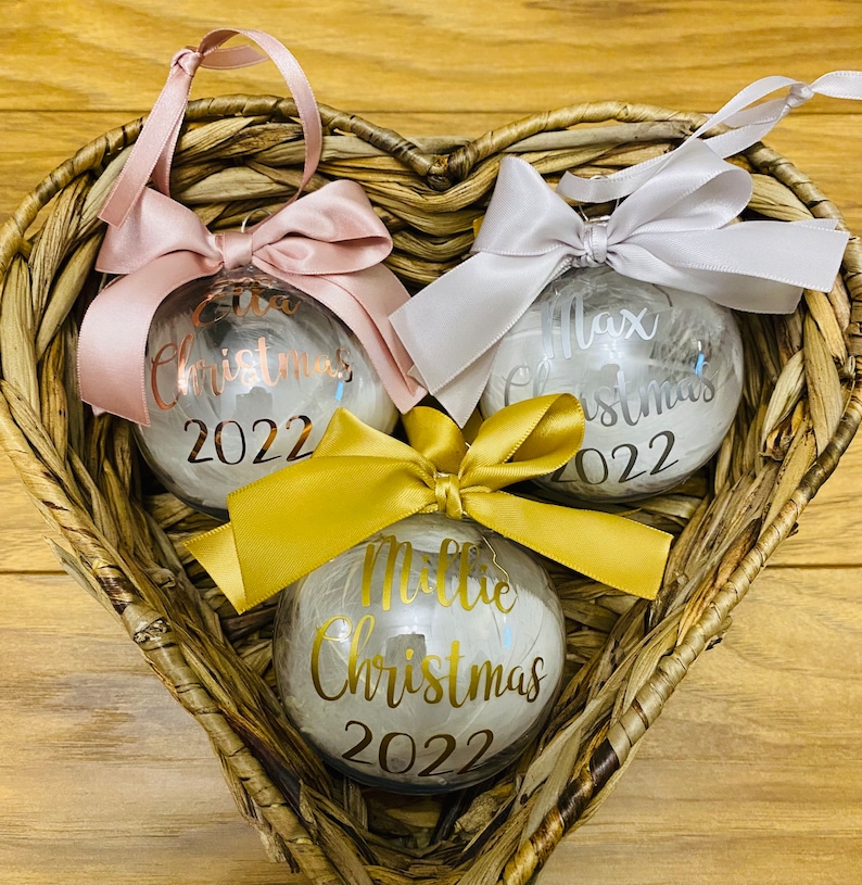 May include: Three clear spherical Christmas baubles filled with white feathers. Each bauble features a satin ribbon bow and custom text in metallic vinyl: Ella Christmas 2022 in rose gold, Max Christmas 2022 in silver, and Millie Christmas 2022 in gold.