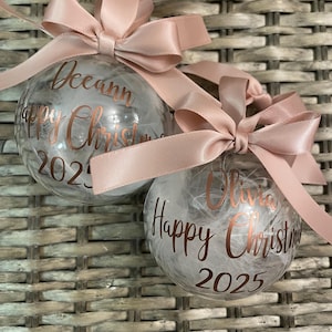 May include: Two clear spherical baubles filled with white feathers, each topped with a dusty pink satin ribbon bow. The baubles feature metallic copper lettering that reads "Deeann Happy Christmas 2025" and "Olivia Happy Christmas 2025" respectively.