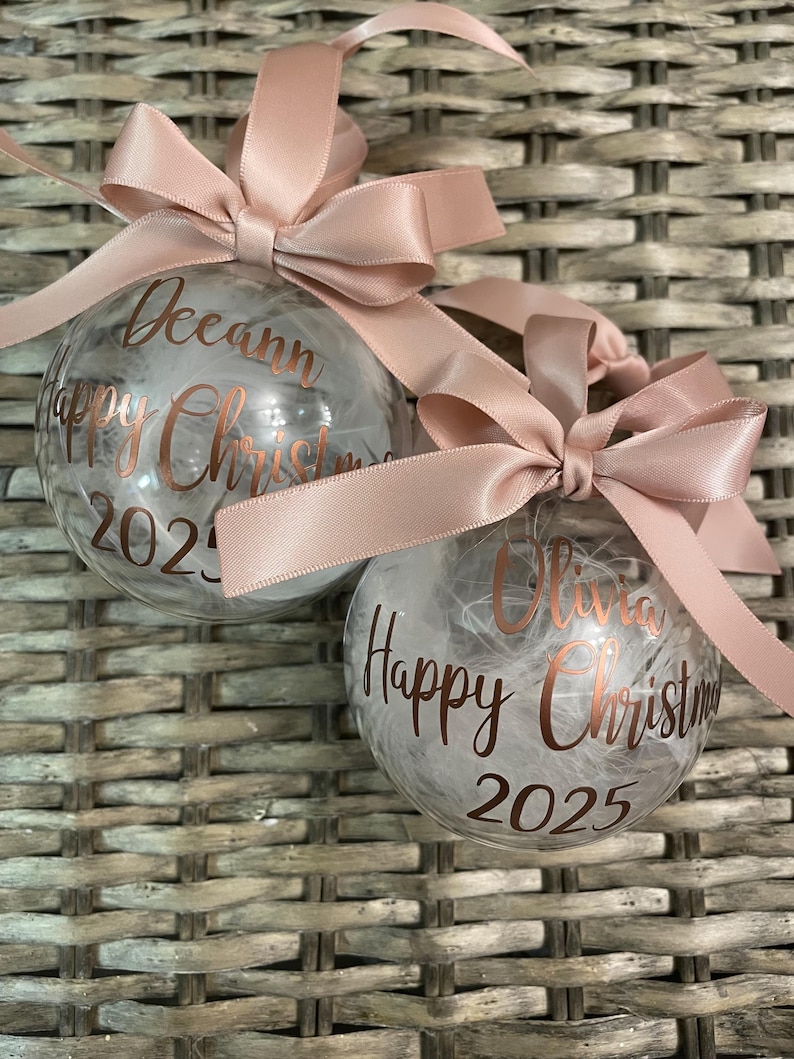May include: Two clear spherical baubles filled with white feathers, each topped with a dusty pink satin ribbon bow. The baubles feature metallic copper lettering that reads "Deeann Happy Christmas 2025" and "Olivia Happy Christmas 2025" respectively.