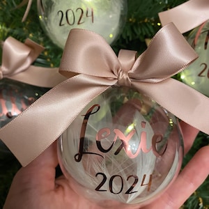 May include: A clear spherical Christmas bauble filled with white feathers, featuring the name Lexie and the year 2024 in metallic rose gold script lettering. The bauble is topped with a decorative satin ribbon bow in a matching rose gold colour.
