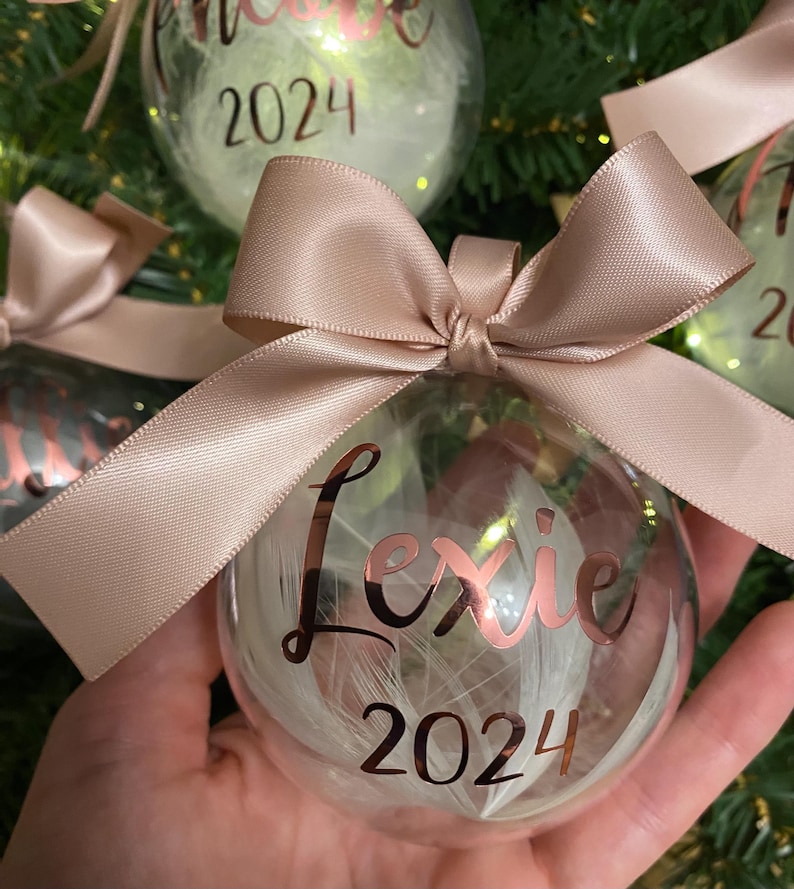 May include: A clear spherical Christmas bauble filled with white feathers, featuring the name Lexie and the year 2024 in metallic rose gold script lettering. The bauble is topped with a decorative satin ribbon bow in a matching rose gold colour.