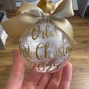 May include: A clear spherical Christmas bauble filled with white feathers, featuring gold cursive text that reads Orla's First Christmas 2025. The bauble is topped with a large, tied gold satin ribbon bow for hanging.