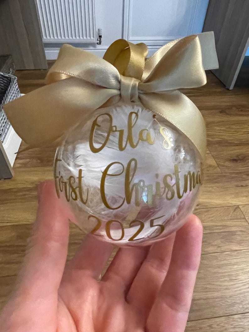 May include: A clear spherical Christmas bauble filled with white feathers, featuring gold cursive text that reads Orla's First Christmas 2025. The bauble is topped with a large, tied gold satin ribbon bow for hanging.