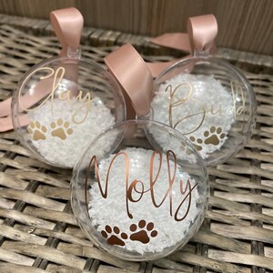 May include: Three clear spherical baubles filled with white faux snow. Each bauble features a name in gold or copper script lettering and two paw prints. The names Clay, Buddy, and Molly are displayed, and each bauble has a pink ribbon loop.