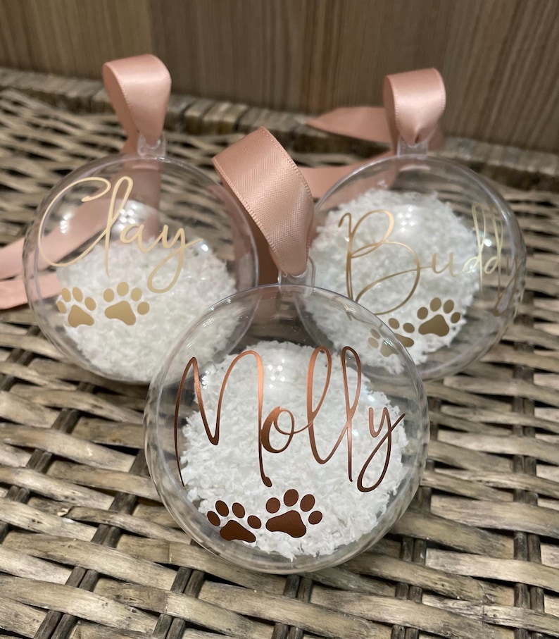 May include: Three clear spherical baubles filled with white faux snow. Each bauble features a name in gold or copper script lettering and two paw prints. The names Clay, Buddy, and Molly are displayed, and each bauble has a pink ribbon loop.