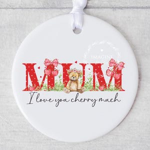 May include: White ceramic bauble featuring a red and white "MUM" design. The letters are adorned with hearts and a small teddy bear wearing a pink flower crown. The text "I love you cherry much" is written beneath the letters.