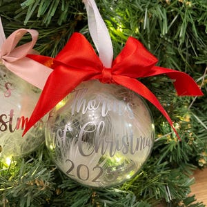 May include: A clear spherical Christmas bauble filled with white feathers, featuring a bright red satin ribbon bow at the top. The bauble is printed with the silver text 'Thorin's first Christmas 2025' and hangs from a white ribbon loop.