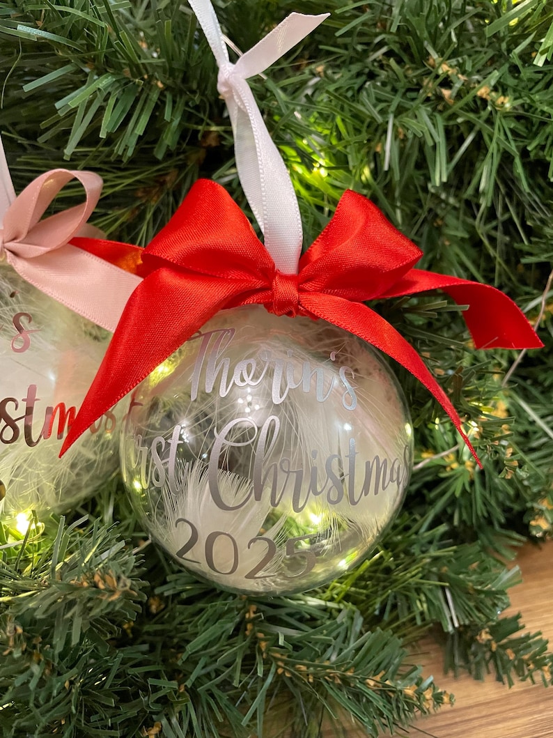 May include: A clear spherical Christmas bauble filled with white feathers, featuring a bright red satin ribbon bow at the top. The bauble is printed with the silver text 'Thorin's first Christmas 2025' and hangs from a white ribbon loop.