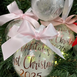 May include: Clear spherical Christmas baubles filled with white feathers and illuminated by internal lights. Each bauble features a pink satin ribbon bow and rose gold lettering that reads Delilah's First Christmas 2025.