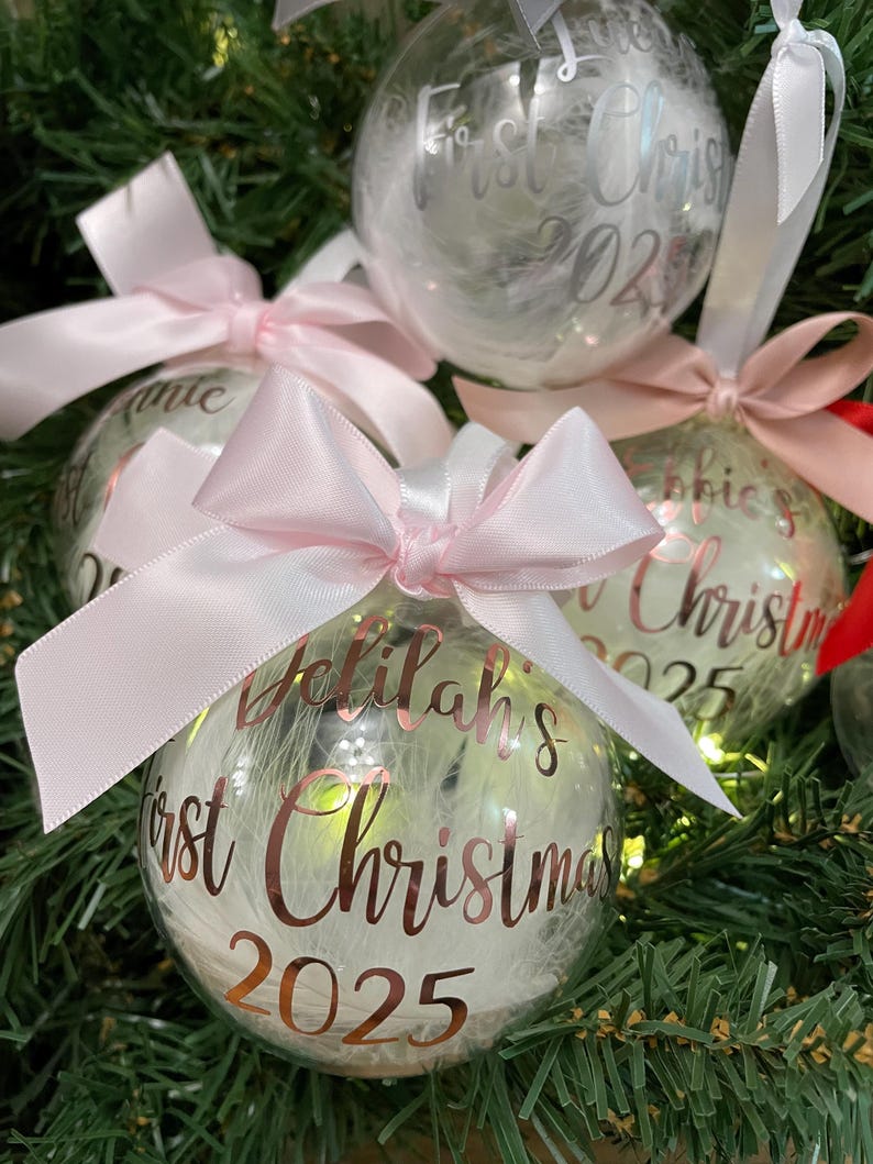 May include: Clear spherical Christmas baubles filled with white feathers and illuminated by internal lights. Each bauble features a pink satin ribbon bow and rose gold lettering that reads Delilah's First Christmas 2025.