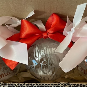 May include: Three clear spherical Christmas baubles filled with white feathers. Each bauble features personalised text in metallic script. The left and right baubles have light pink satin bows, while the centre bauble has a bright red satin bow.