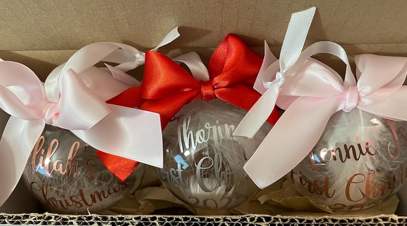 May include: Three clear spherical Christmas baubles filled with white feathers. Each bauble features personalised text in metallic script. The left and right baubles have light pink satin bows, while the centre bauble has a bright red satin bow.