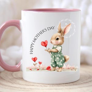 May include: White ceramic mug with a pink rim and handle. The mug features a cartoon bunny holding a red heart flower and the text "Happy Mother's Day".