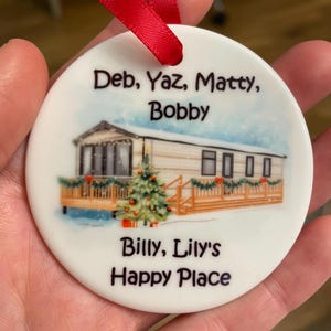 May include: A round white ceramic ornament with a red ribbon hanger. The centre features a printed illustration of a mobile home with a Christmas tree and garland. The text reads: Deb, Yaz, Matty, Bobby, Billy, Lily's Happy Place.