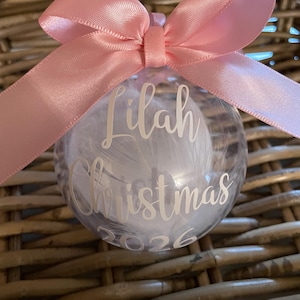 May include: A clear spherical ornament filled with white feathers, featuring a pink satin ribbon bow at the top. The front is personalised with white text reading Lilah Christmas 2026 in a script and sans-serif font combination.