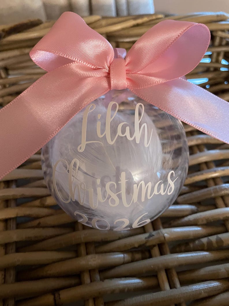 May include: A clear spherical ornament filled with white feathers, featuring a pink satin ribbon bow at the top. The front is personalised with white text reading Lilah Christmas 2026 in a script and sans-serif font combination.