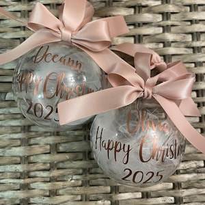 May include: Two clear spherical Christmas baubles filled with white feathers. Each bauble features rose gold cursive text reading "Happy Christmas" with a name, "Deeann" or "Olivia," and the year "2025." A dusty pink satin ribbon bow is tied at the top.