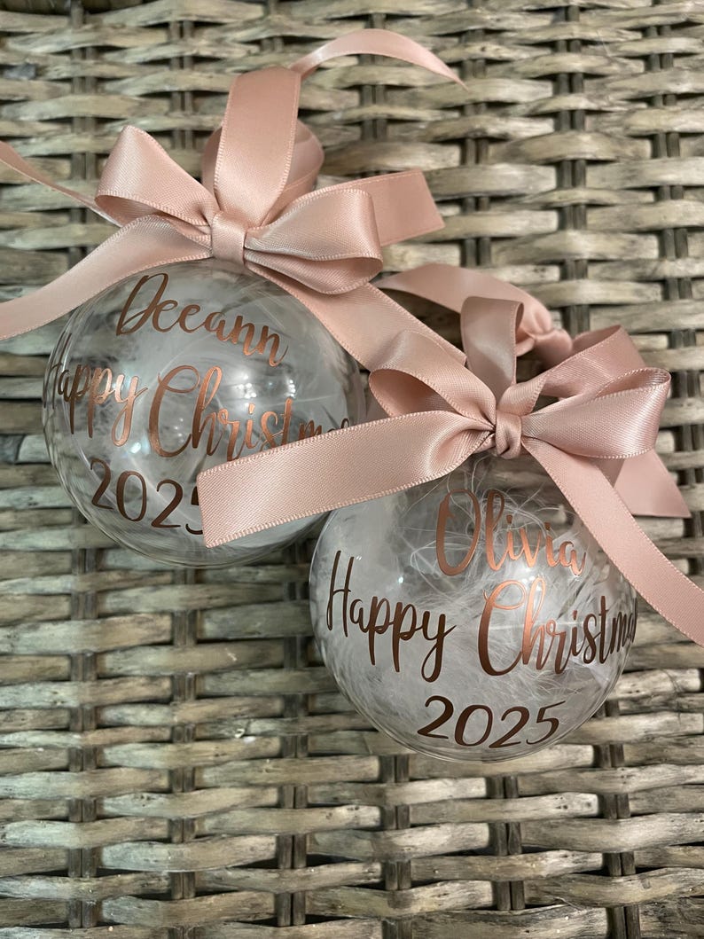 May include: Two clear spherical Christmas baubles filled with white feathers. Each bauble features rose gold cursive text reading "Happy Christmas" with a name, "Deeann" or "Olivia," and the year "2025." A dusty pink satin ribbon bow is tied at the top.