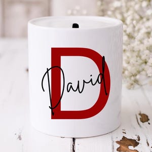 May include: White ceramic money bank featuring a red letter "D" and the name "David" in black script. The money bank has a slot on top for inserting coins or notes. The design is simple and elegant.