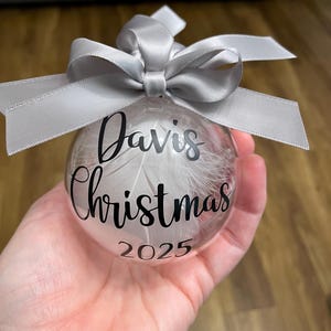 May include: A clear spherical bauble filled with white feathers, featuring the black text "Davis Christmas 2025" printed on the front. The bauble is topped with a decorative silver satin ribbon tied into a bow at the top attachment point.