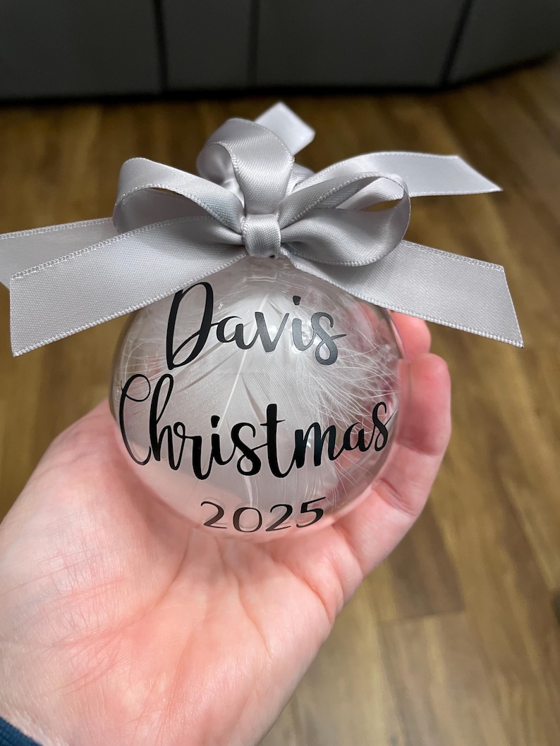 May include: A clear spherical bauble filled with white feathers, featuring the black text "Davis Christmas 2025" printed on the front. The bauble is topped with a decorative silver satin ribbon tied into a bow at the top attachment point.