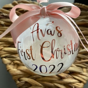 May include: A clear spherical Christmas bauble filled with white feathers, featuring rose gold lettering that reads Ava's First Christmas 2022. The bauble is topped with a light pink satin ribbon tied into a bow for hanging purposes.