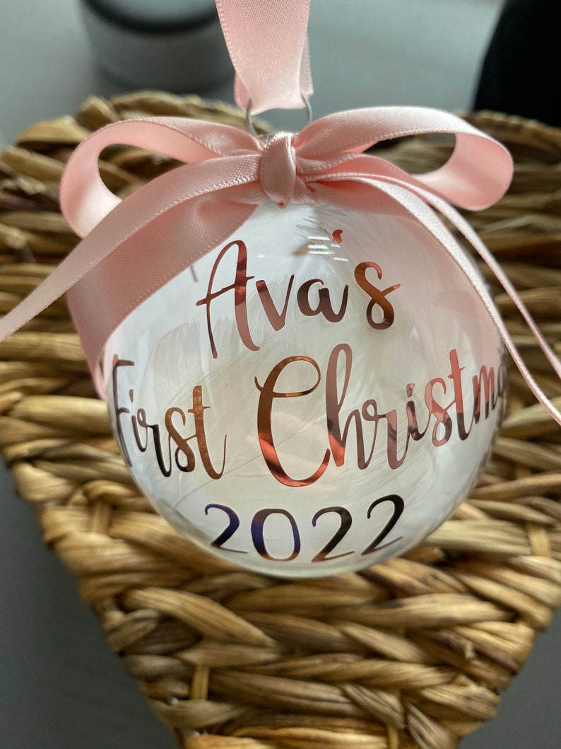 May include: A clear spherical Christmas bauble filled with white feathers, featuring rose gold lettering that reads Ava's First Christmas 2022. The bauble is topped with a light pink satin ribbon tied into a bow for hanging purposes.